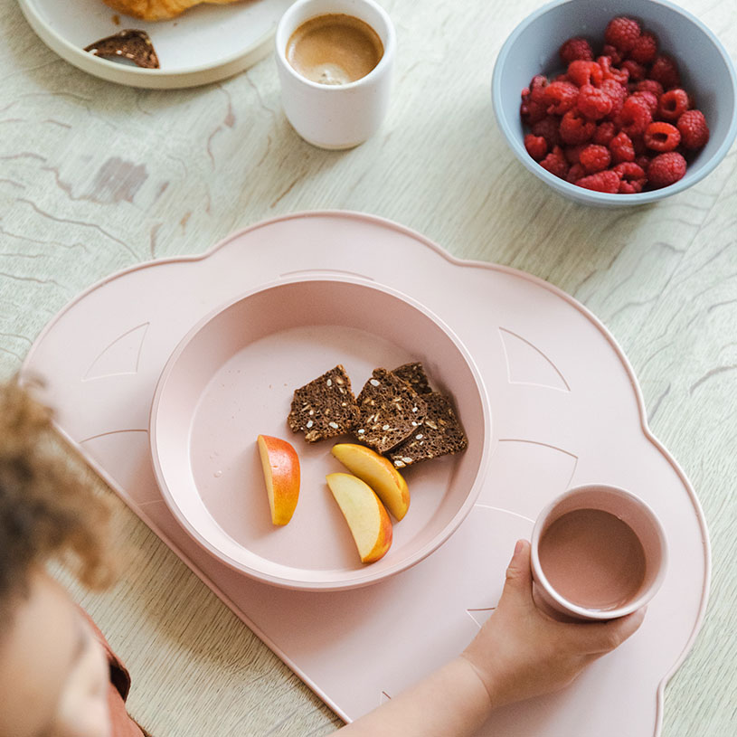 Barnhand med rosa mugg, bröd och äppelbitar på rosa tallrik, hallon i skål.
