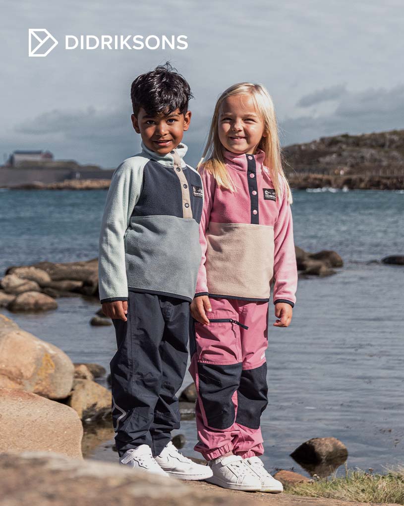 Två barn klättrar på ett räcke vid havet, iförda Didriksons kläder.