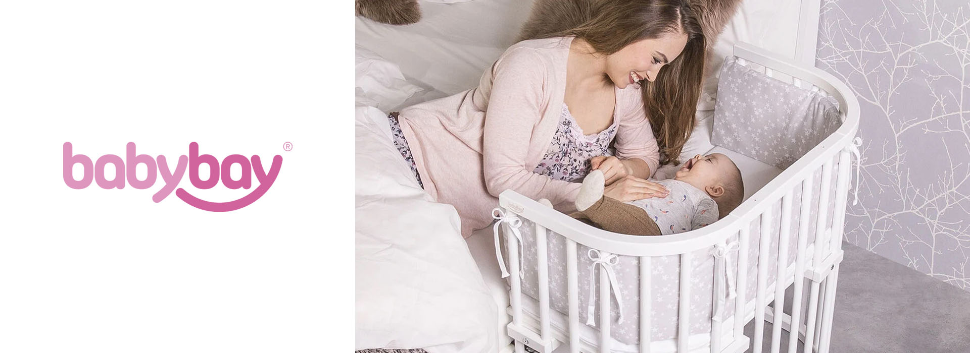 Mamma skrattar med bebis i grå/vit spjälsäng vid sängkant.