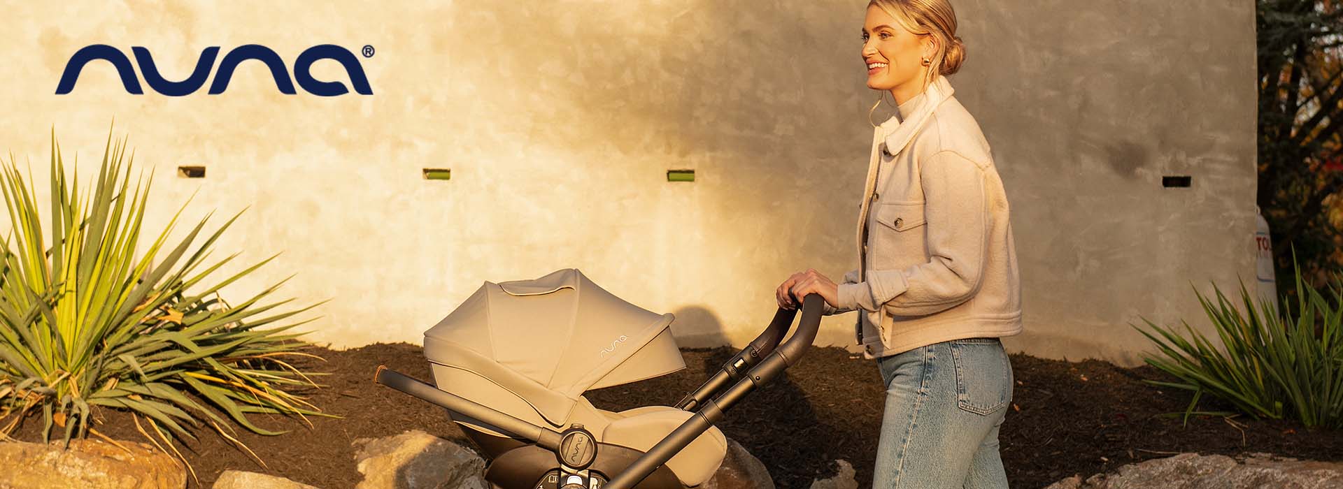 Kvinna i jacka promenerar med barnvagn i solsken.