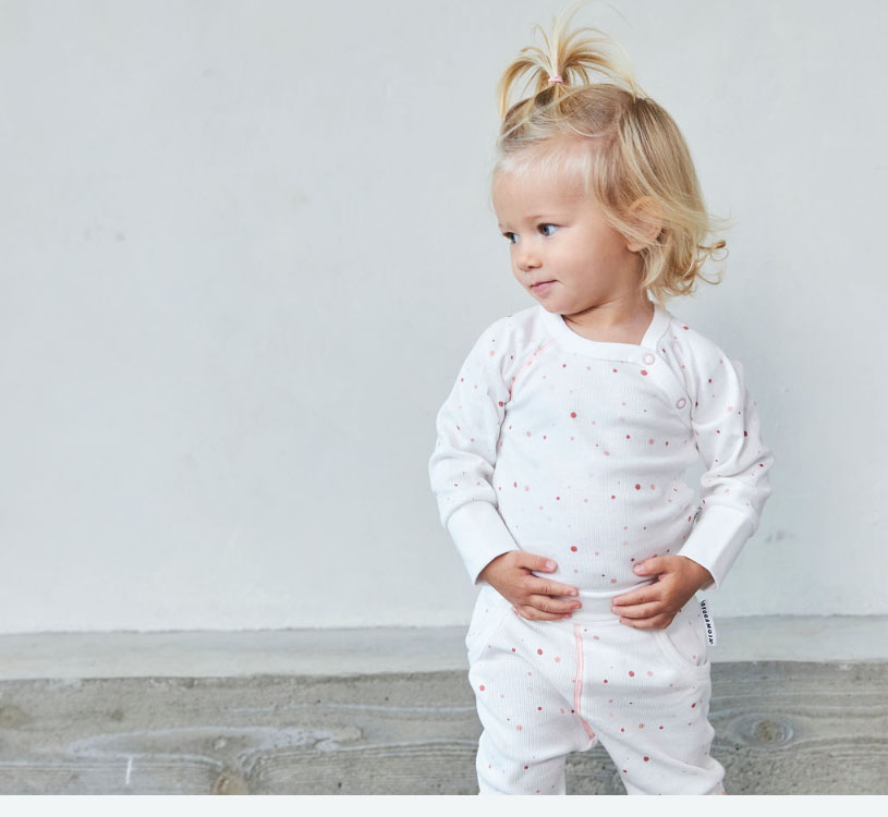 Litet barn i vit pyjamas med prickar, står mot en grå bakgrund.