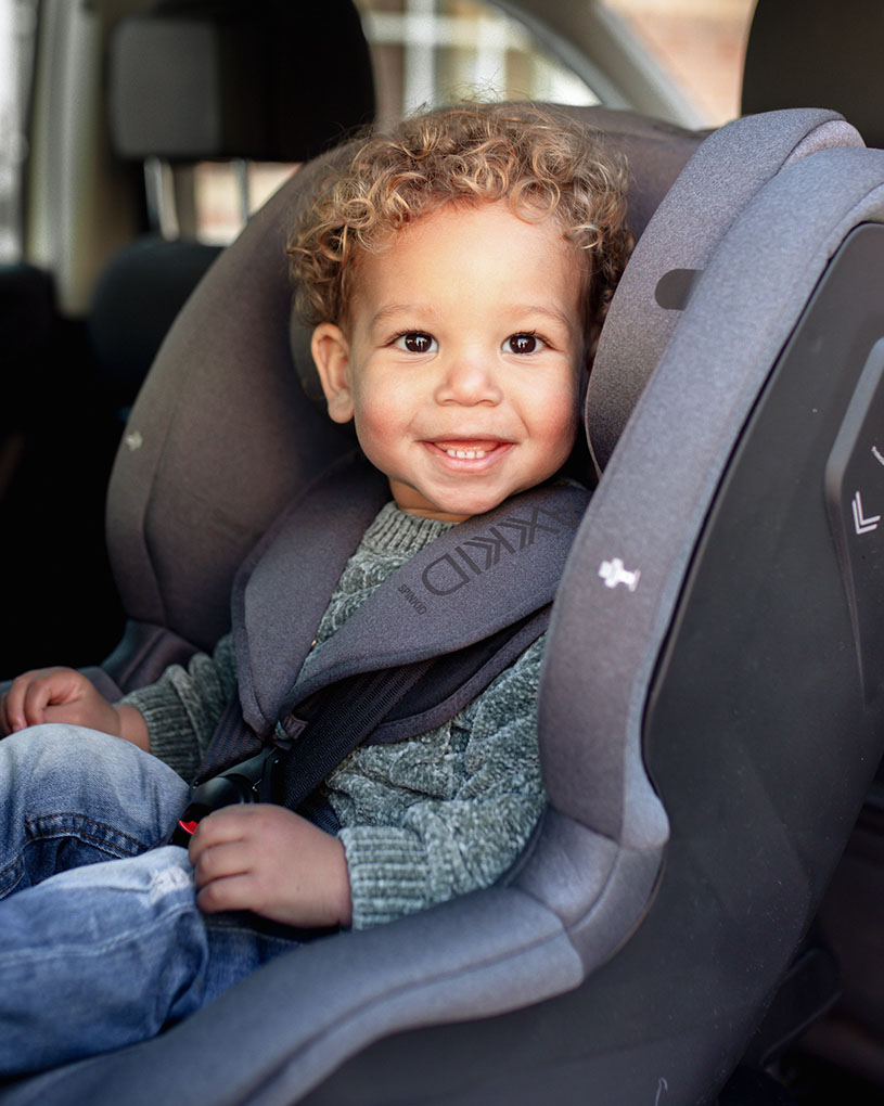 Barn med lockigt hår ler och sitter i en bilbarnstol.