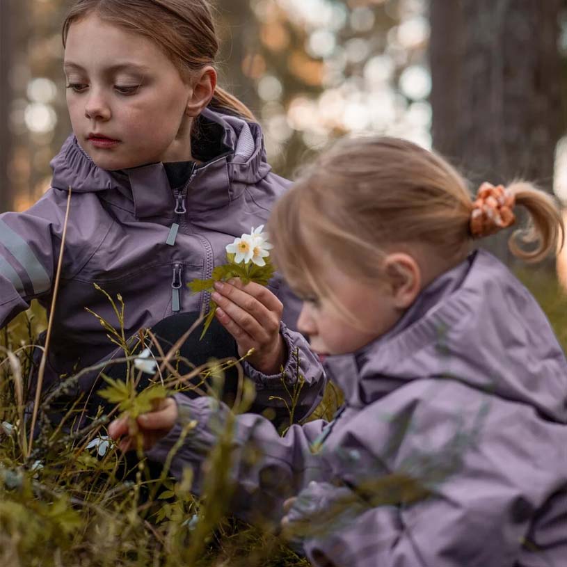 Två barn i färgglada jackor står vid ett träd och ler.