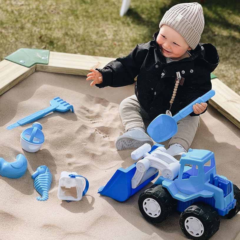 Barn leker i sandlådan med leksaker, inklusive en blå spade och en traktor.