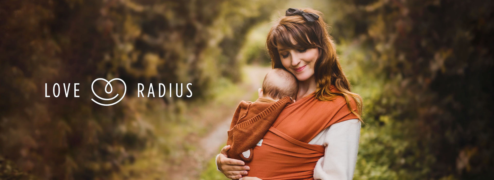 Kvinna håller bebis i sjal i skogsområde, logotyp till vänster.