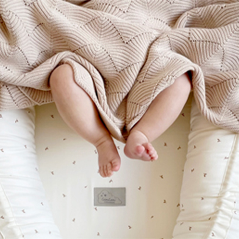 Bebisfötter under en beige filt på en vit madrass.