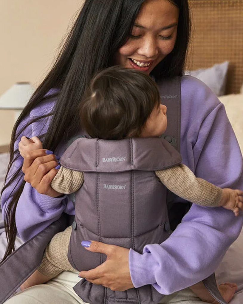 Kvinna i lila tröja bär bebis i bärsele inomhus.