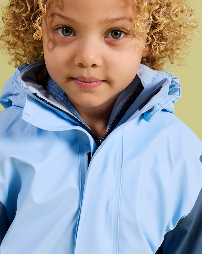 Barn med lockigt hår i en ljusblå regnjacka med texten 'Fodrade regnställ'.