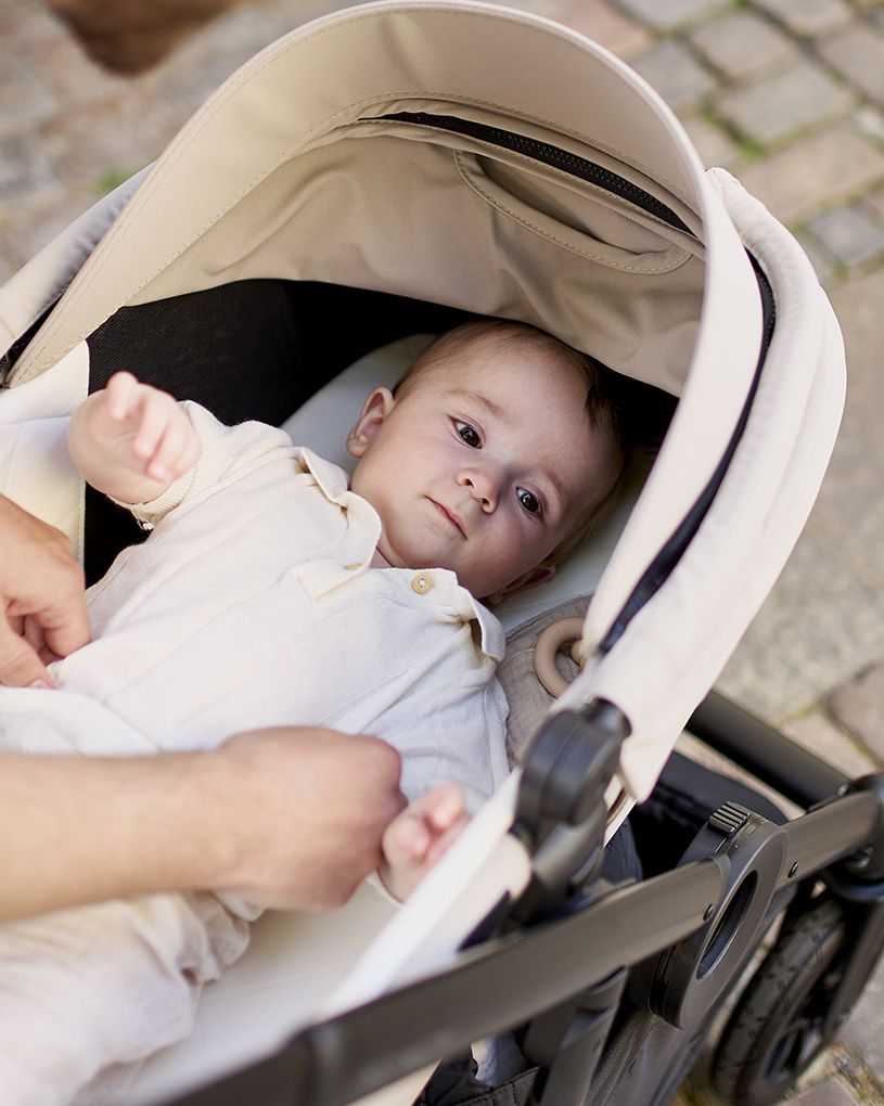 En svart barnvagn med silverram och brunt handtag står på ett ljust trägolv.