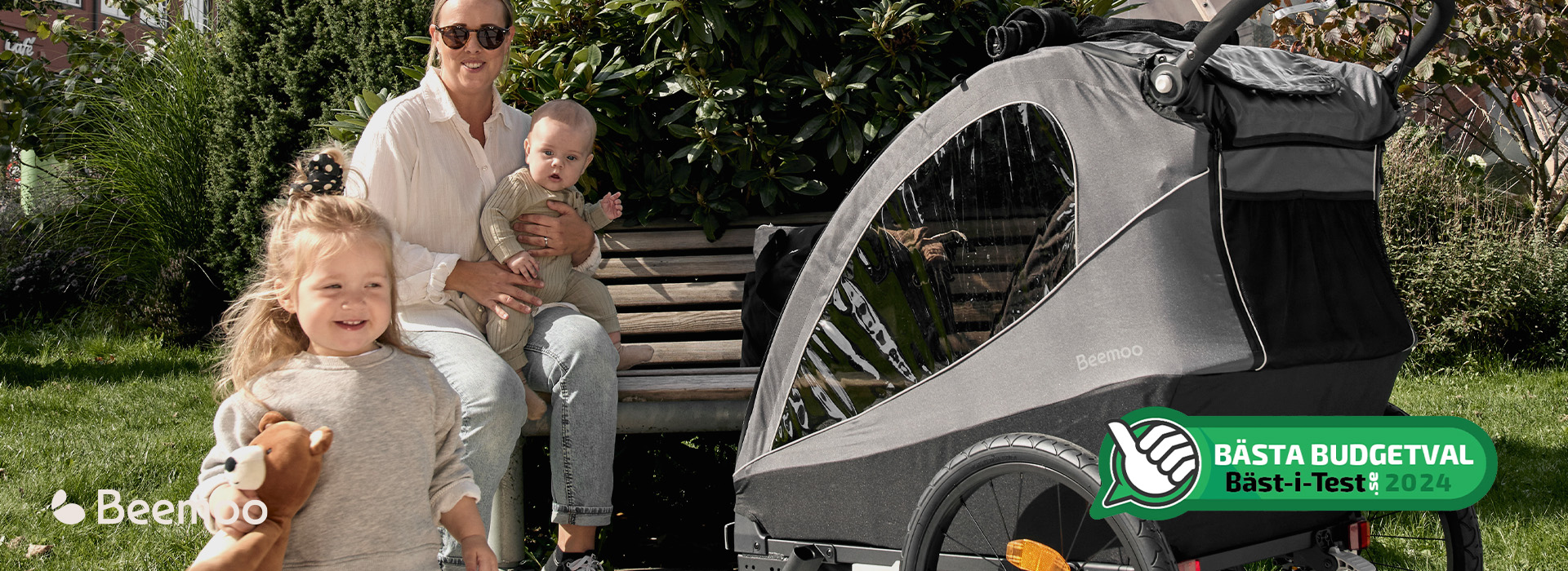 Kvinna och två barn vid parkbänk med cykelvagn från Beemoo, leende i solsken.
