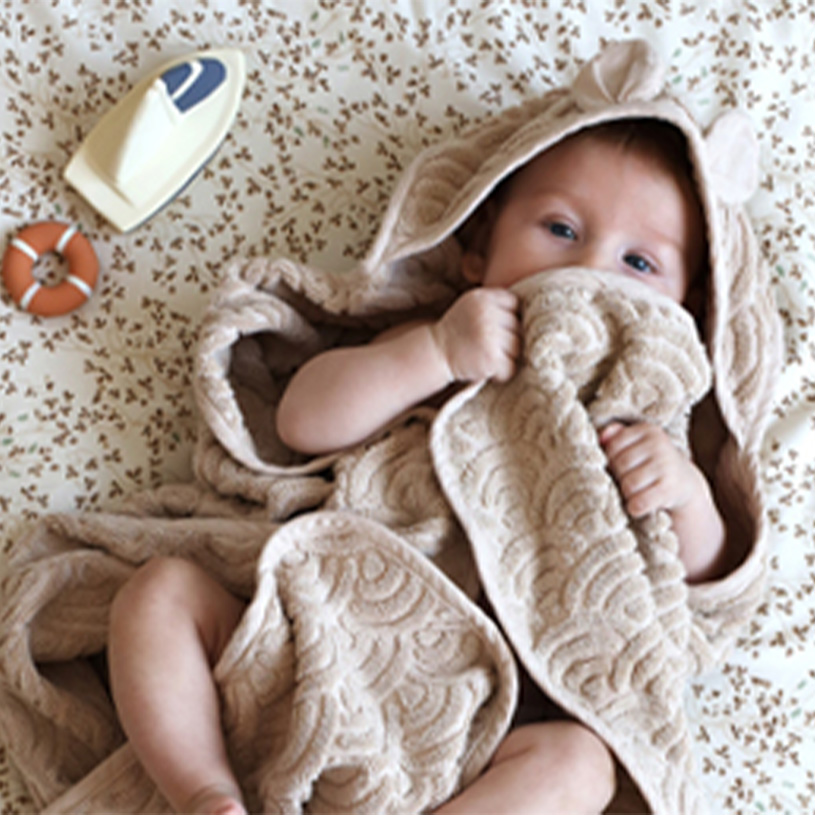 Baby insvept i en beige filt med öronhuva, leksaksbåt och livboj på mönstrad filt.