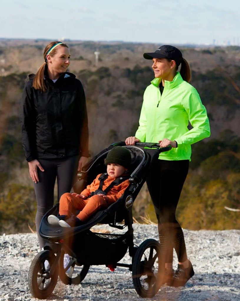 Två kvinnor med en barnvagn på en grusväg med utsikt över en skog.