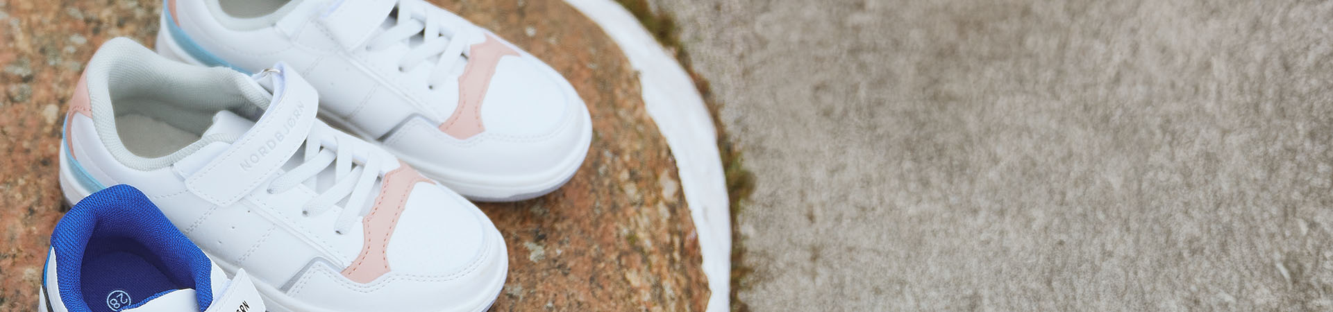 Två vita barnskor med rosa detaljer på stenig underlag.