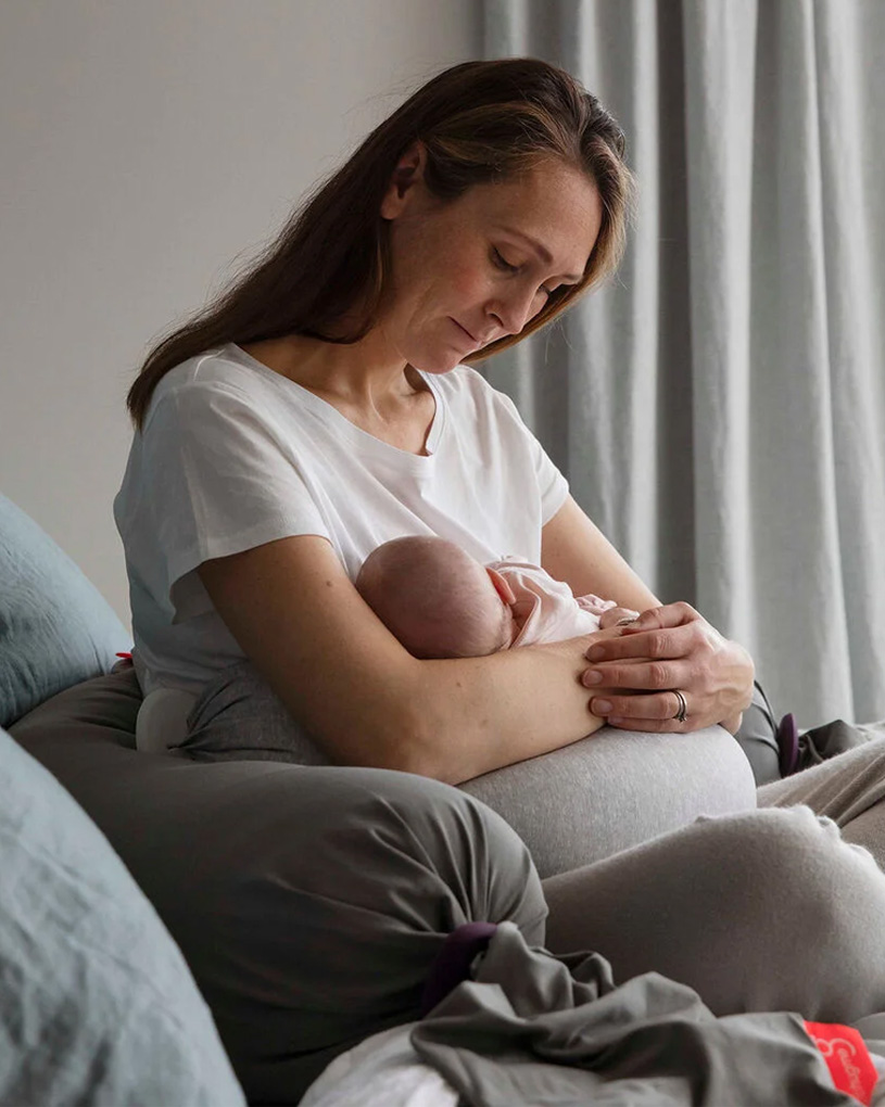Kvinna i vit T-shirt håller en bebis i famnen, sittande i en grå soffa.