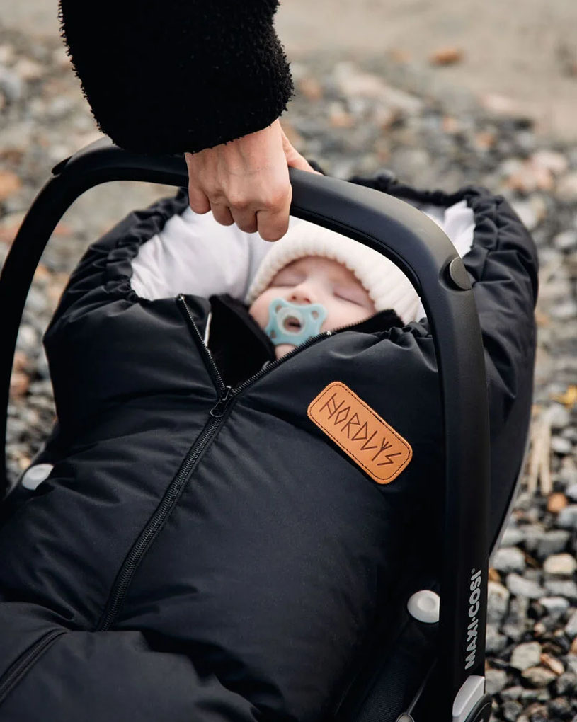Bebis i svart sovsäck i bilbarnstol med napp, vuxen hand håller handtaget.
