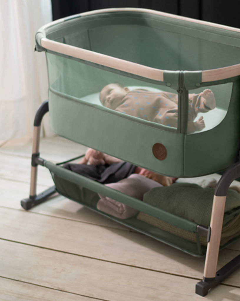 Spädbarn sover i en grön barnsäng med förvaringshylla under.