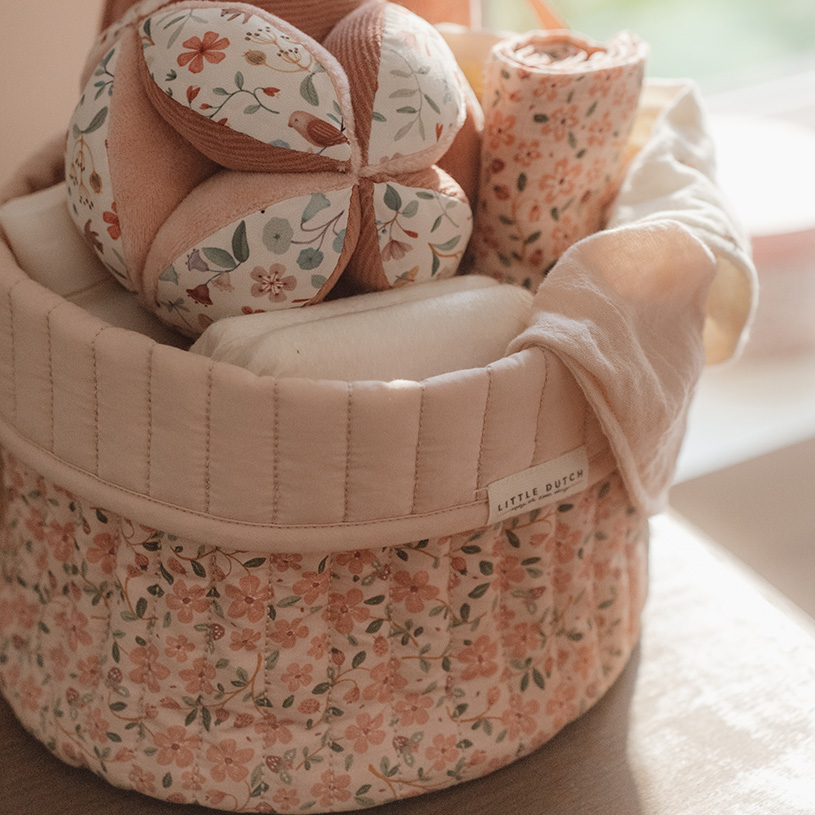 Rund korg med blommigt tyg med babyprodukter inuti, inkl. leksaksboll och filt.