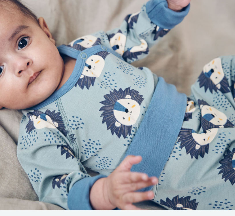 Bebis i blå pyjamas med lejonmönster, liggande på beige kudde.